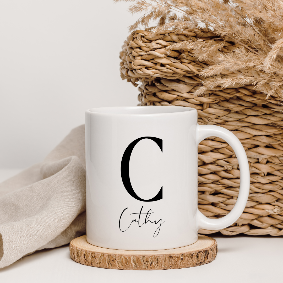 White mug with a black letter 'C' and name on a wooden coaster, with pampas grass and woven basket in the background.